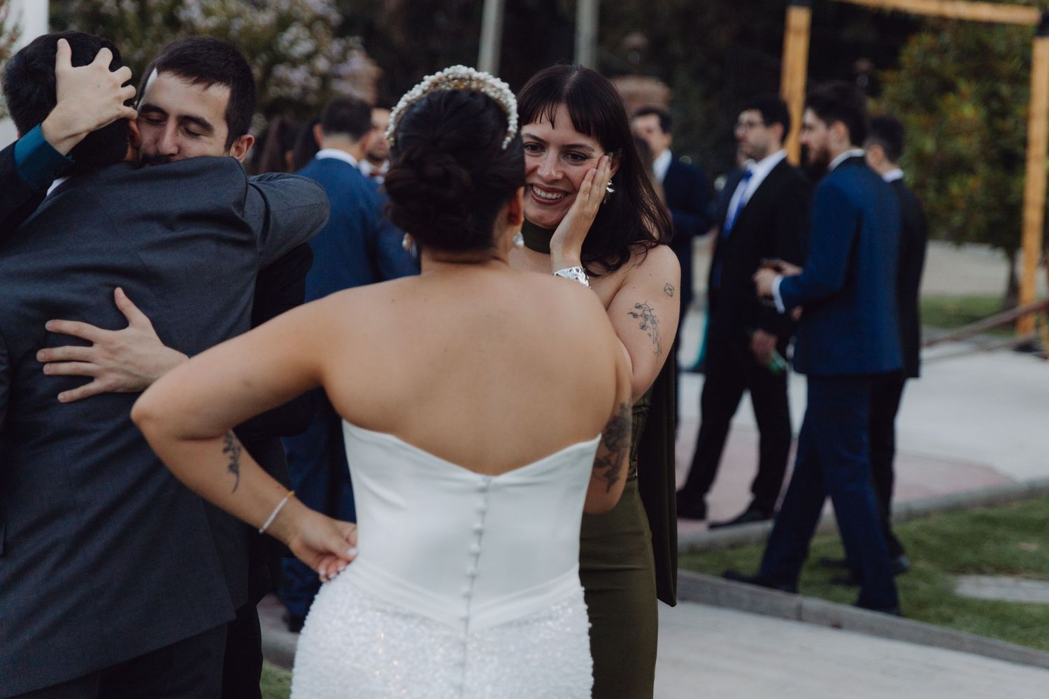 Bride in strapless white gown smiles emotionally while guests embrace at an outdoor wedding.