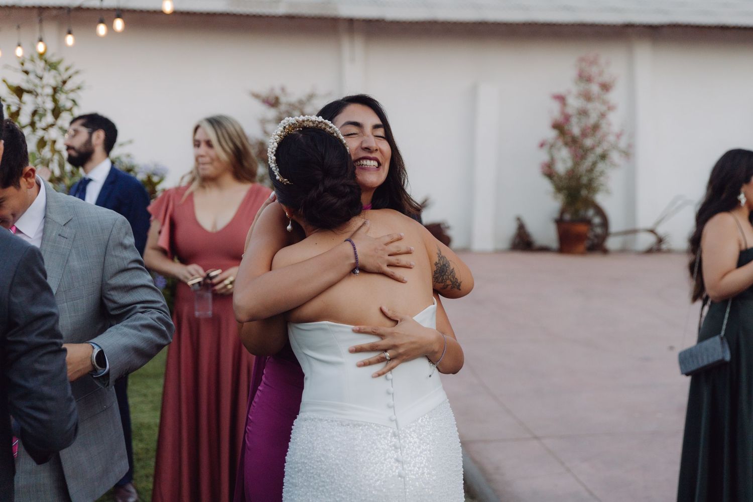 Smiling wedding guest embraces the bride in a white lace gown at an outdoor reception.