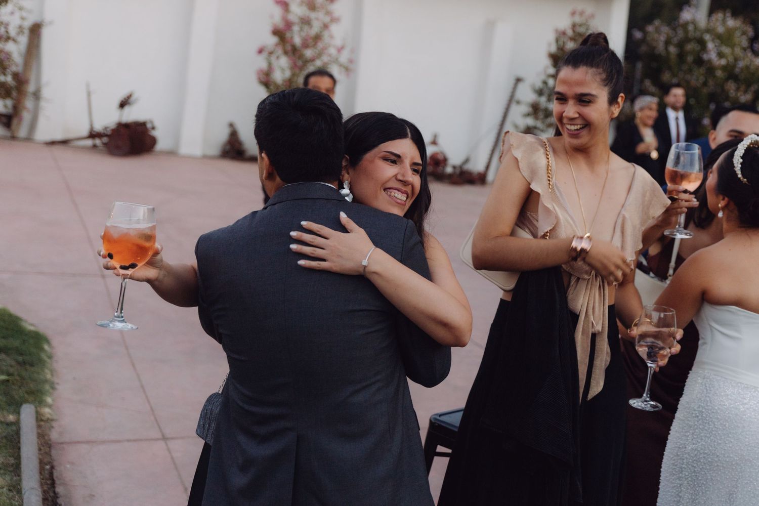 Wedding guest in black outfit hugs an attendee while others hold drinks at an outdoor reception.