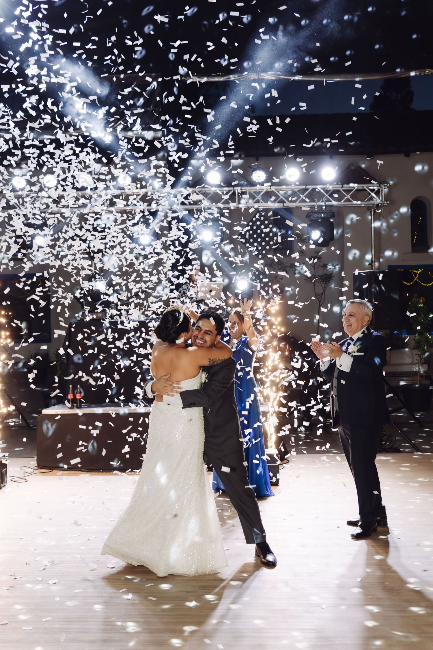 Newlyweds slow dance together surrounded by cascading white confetti and bright stage lighting at their wedding.