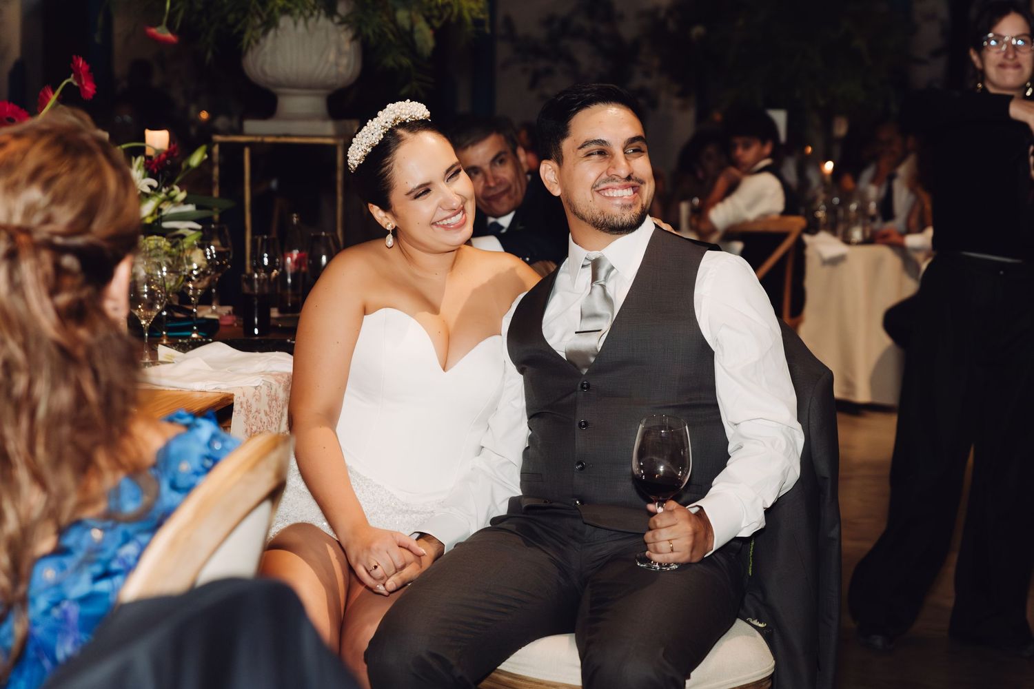 Happy bride and groom smile at guests while seated at their wedding reception table with wine.