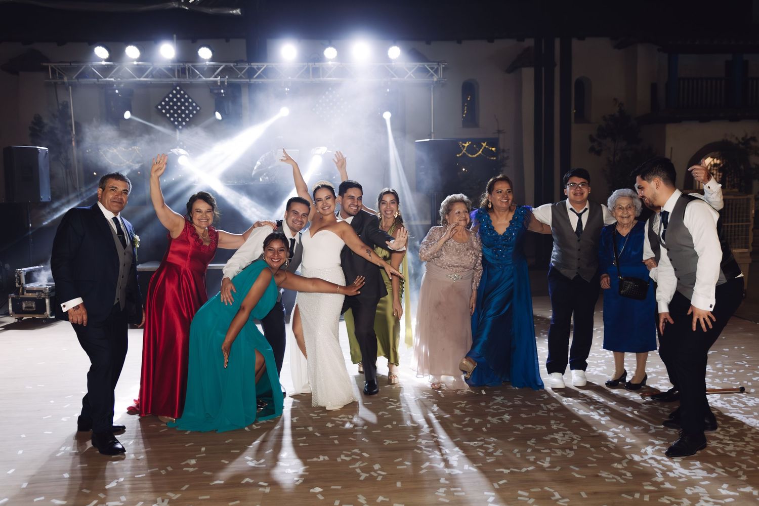 Wedding party group photo with bride and groom surrounded by family and friends celebrating on a dance floor with stage lighting.