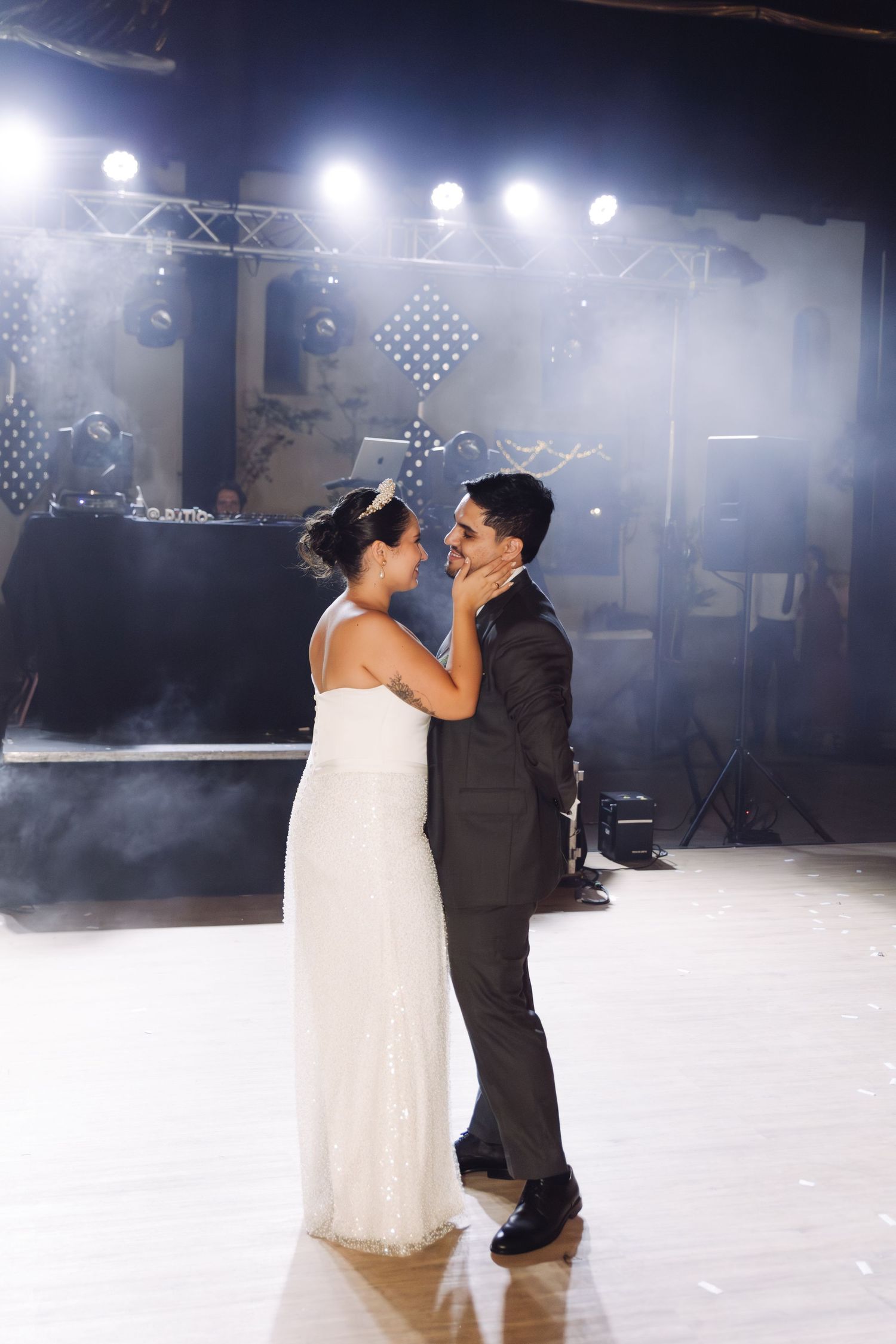 Bride tenderly touches grooms face during their first dance in a beautifully lit wedding venue with fog effects.
