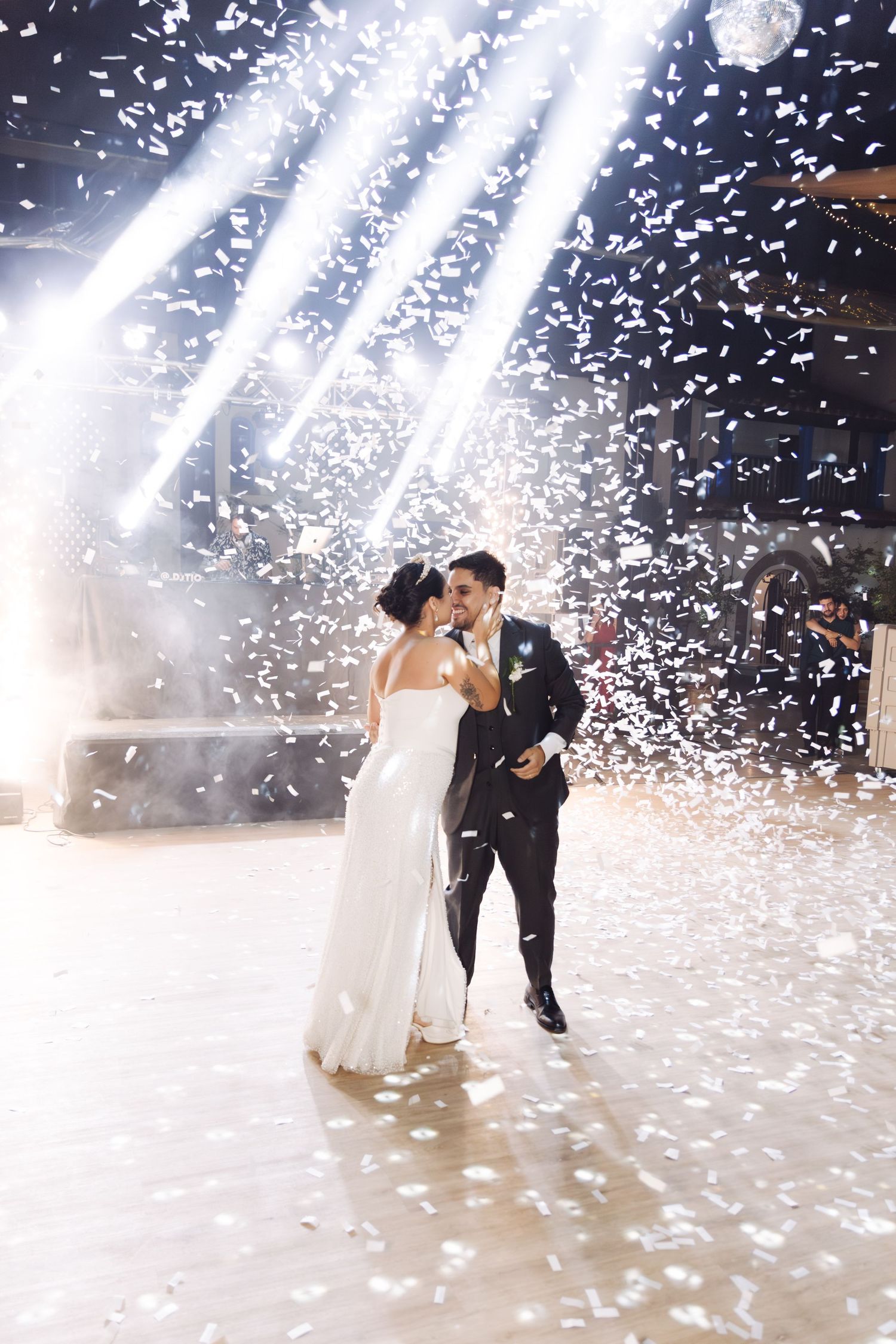 Bride and groom share a romantic kiss on the dance floor as white confetti rains down under bright spotlights.