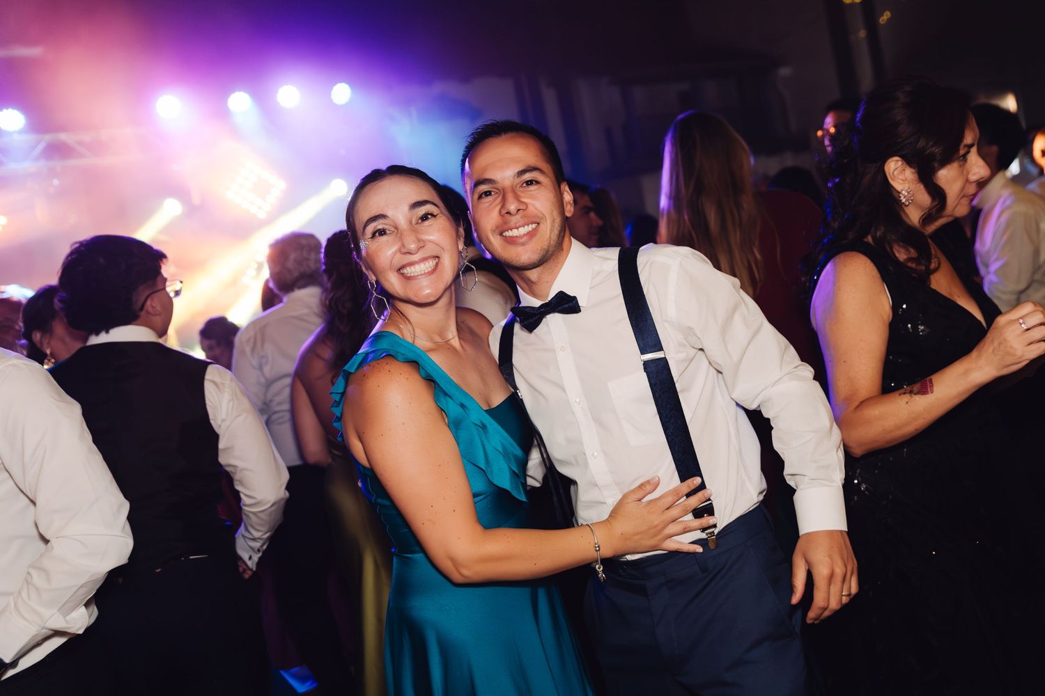 Smiling couple poses together at a vibrant wedding reception with colorful lights and dancing guests behind them.