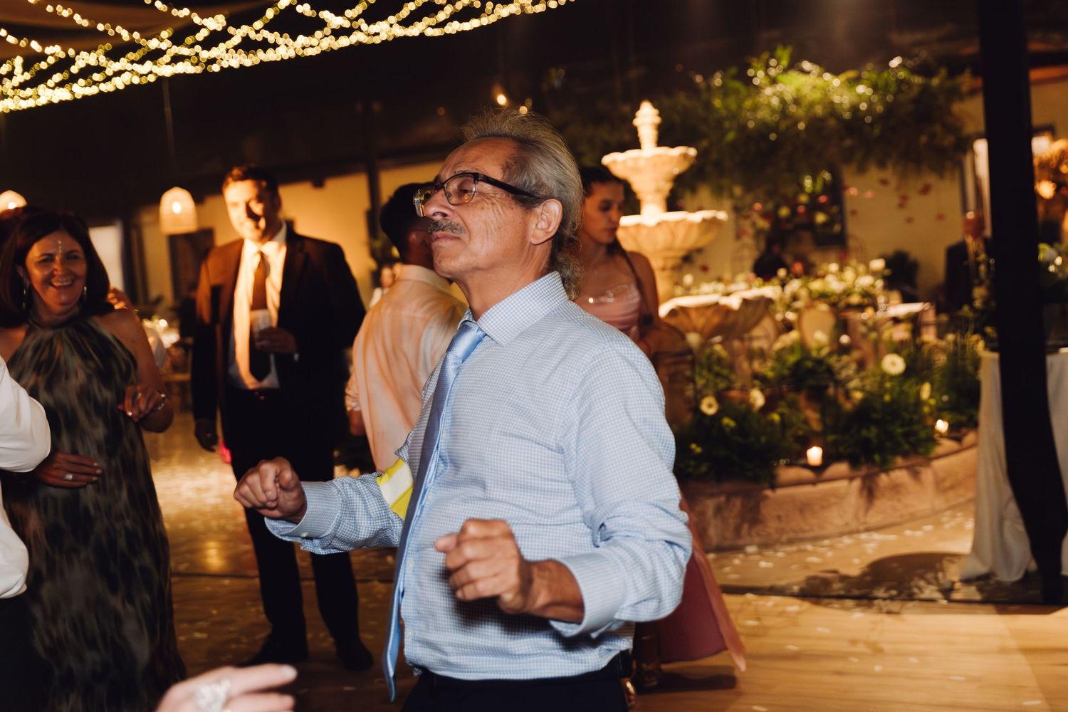 Man dancing outdoors at a wedding reception with a fountain and string lights in the background.