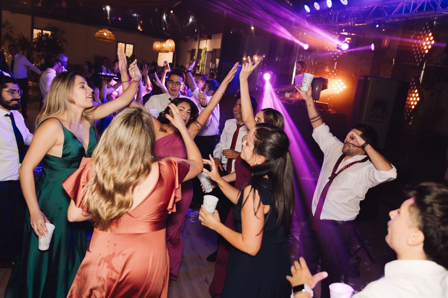 Excited wedding guests dancing with arms raised under purple lights at a festive reception.