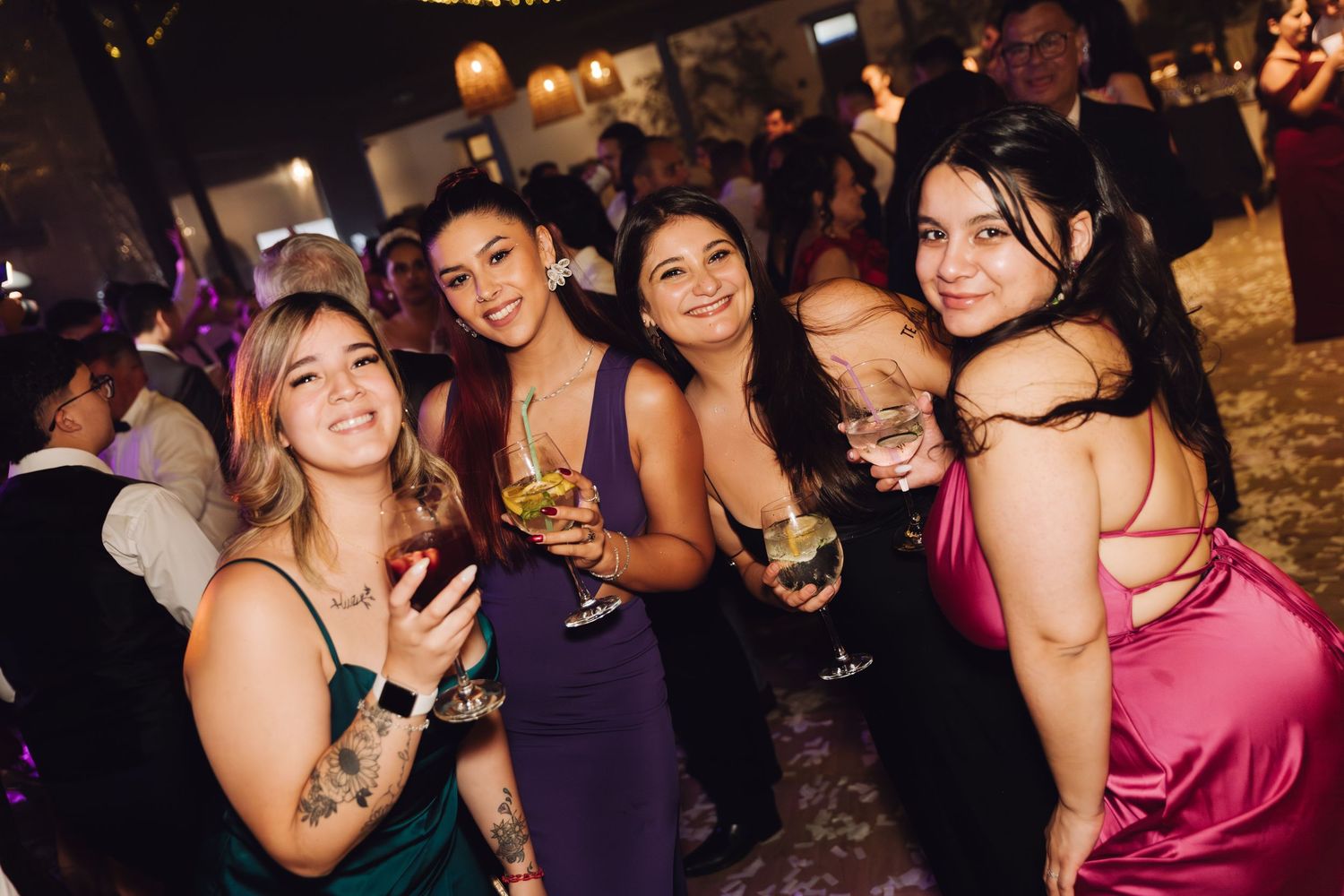 Four smiling women in formal gowns holding cocktails and posing at a wedding reception.