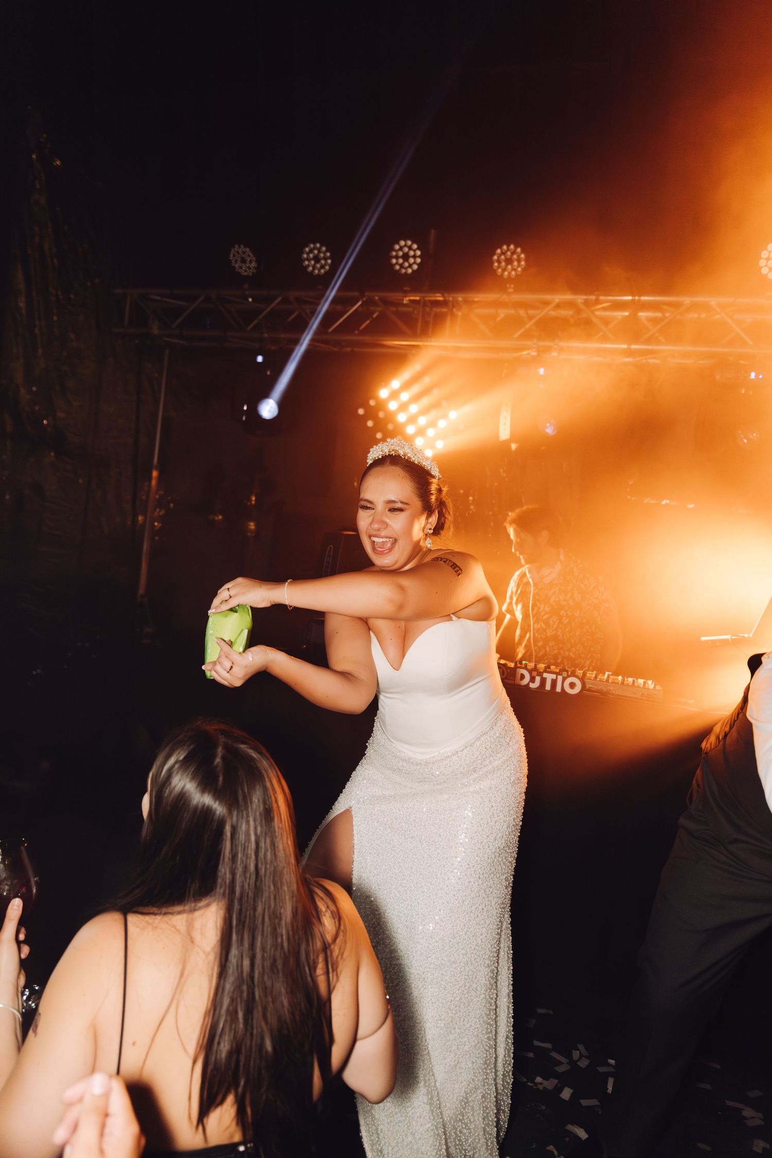 Bride in white gown laughing and dancing with guests under dramatic stage lighting.