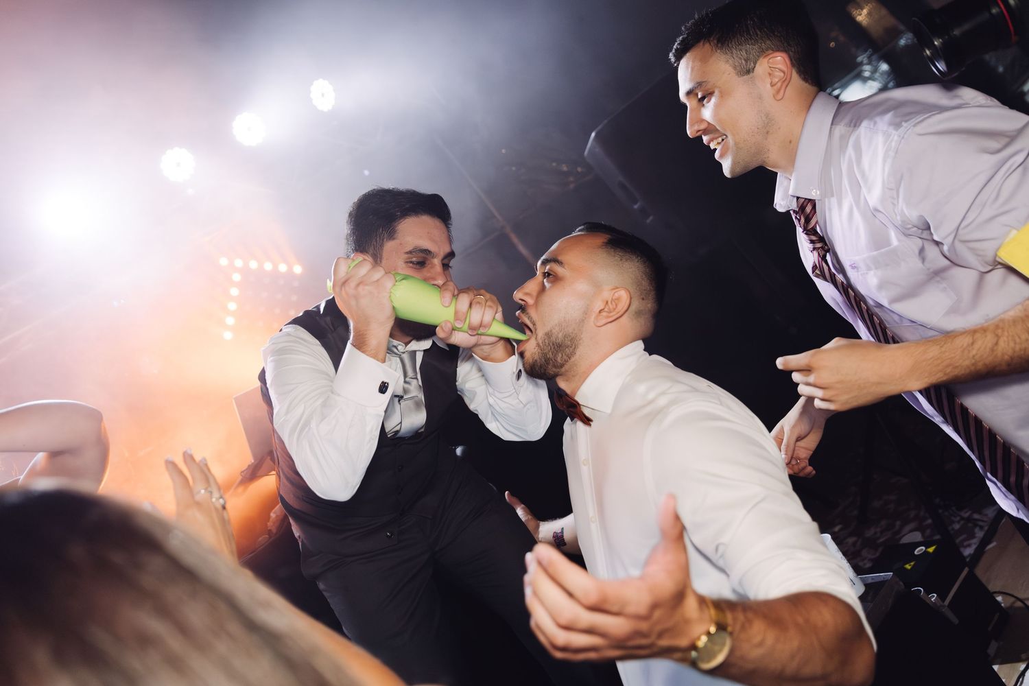 Men drinking from green bottles together under bright lights at a wedding celebration.