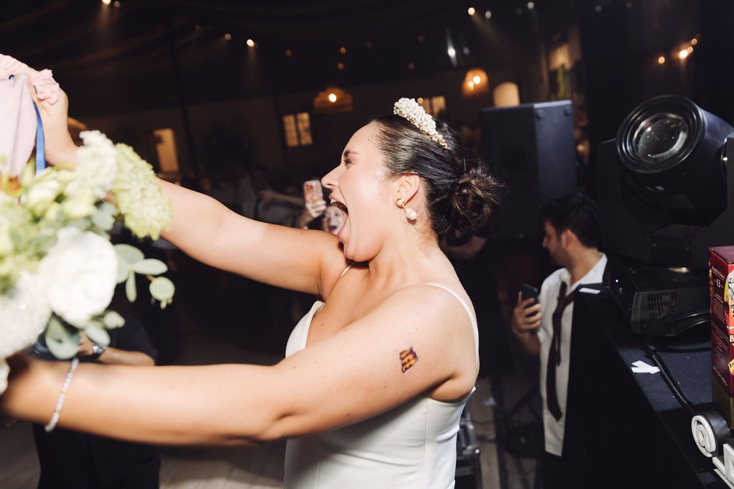 Excited bride in white dress reaching for bouquet at a festive wedding reception.