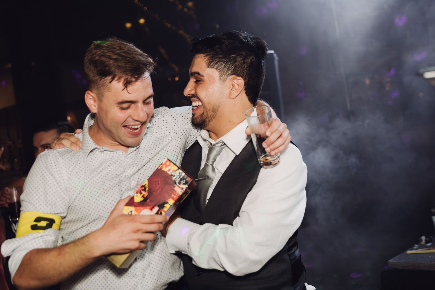 Two men smiling and embracing warmly under moody lighting at a wedding celebration.