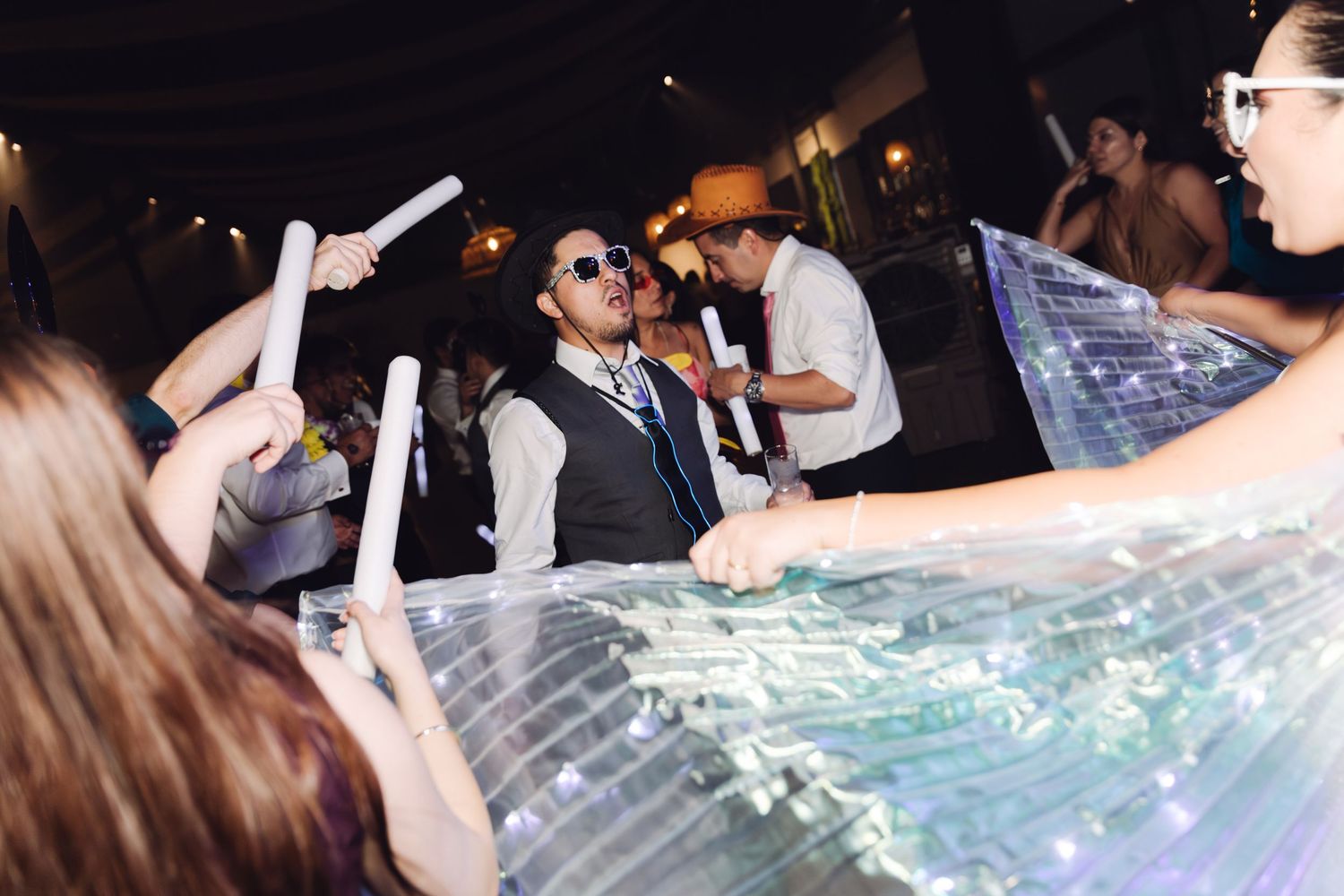 Groom in vest and sunglasses dancing with guests holding glow sticks at wedding reception.