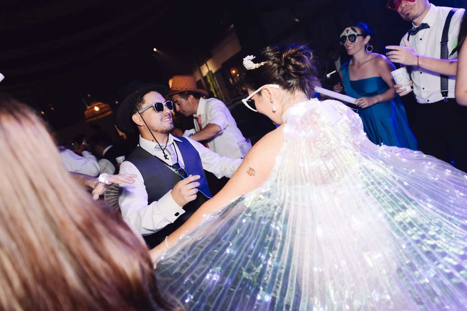 Bride in light-up cape and groom in sunglasses dancing at festive wedding reception.