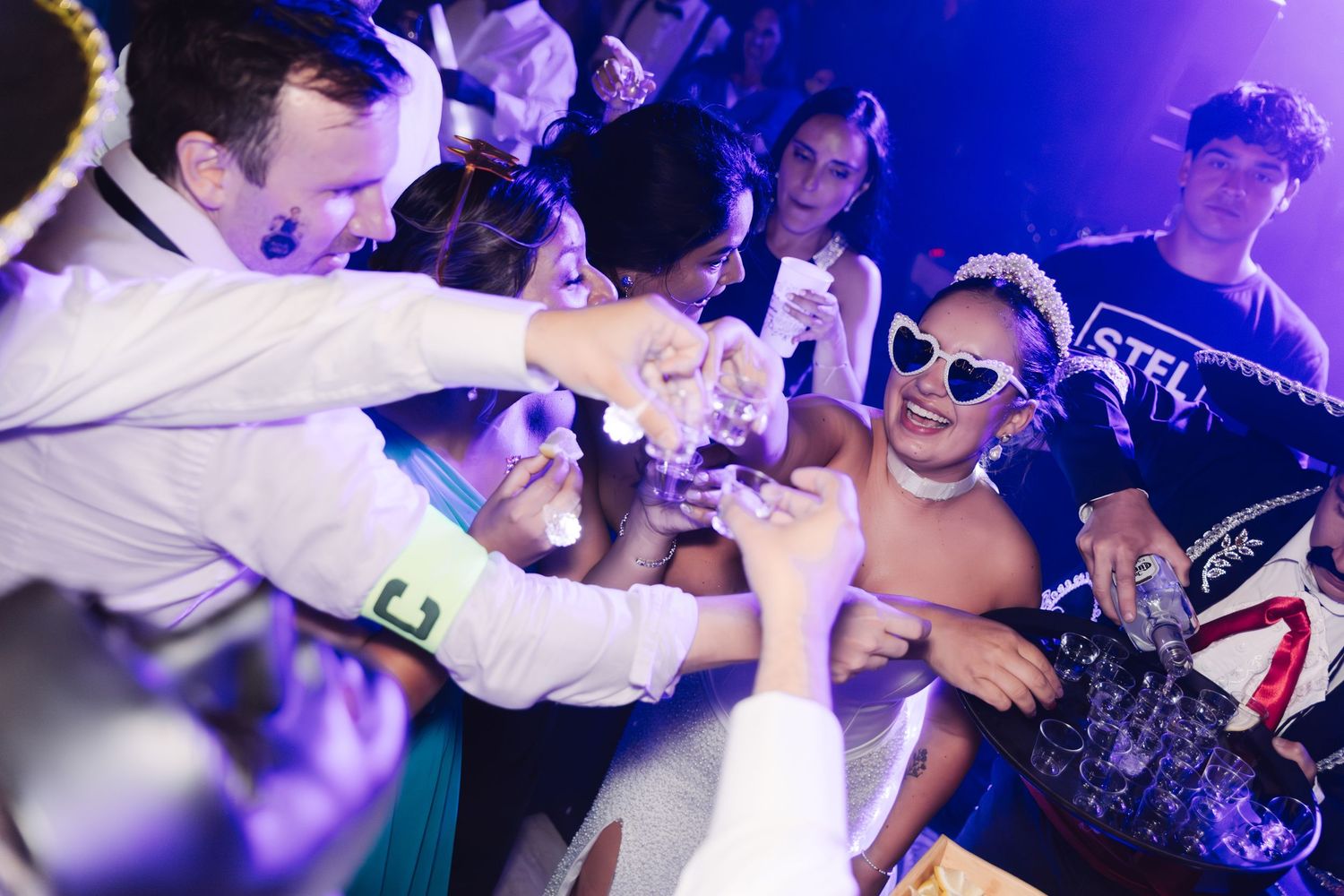 Wedding guests toasting drinks together under purple lights at lively reception party.