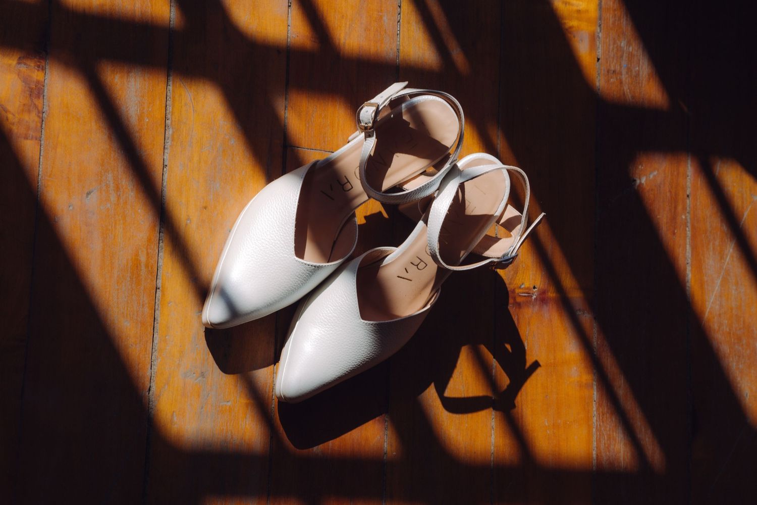 White bridal heeled sandals on wooden floor with dramatic shadow light pattern.