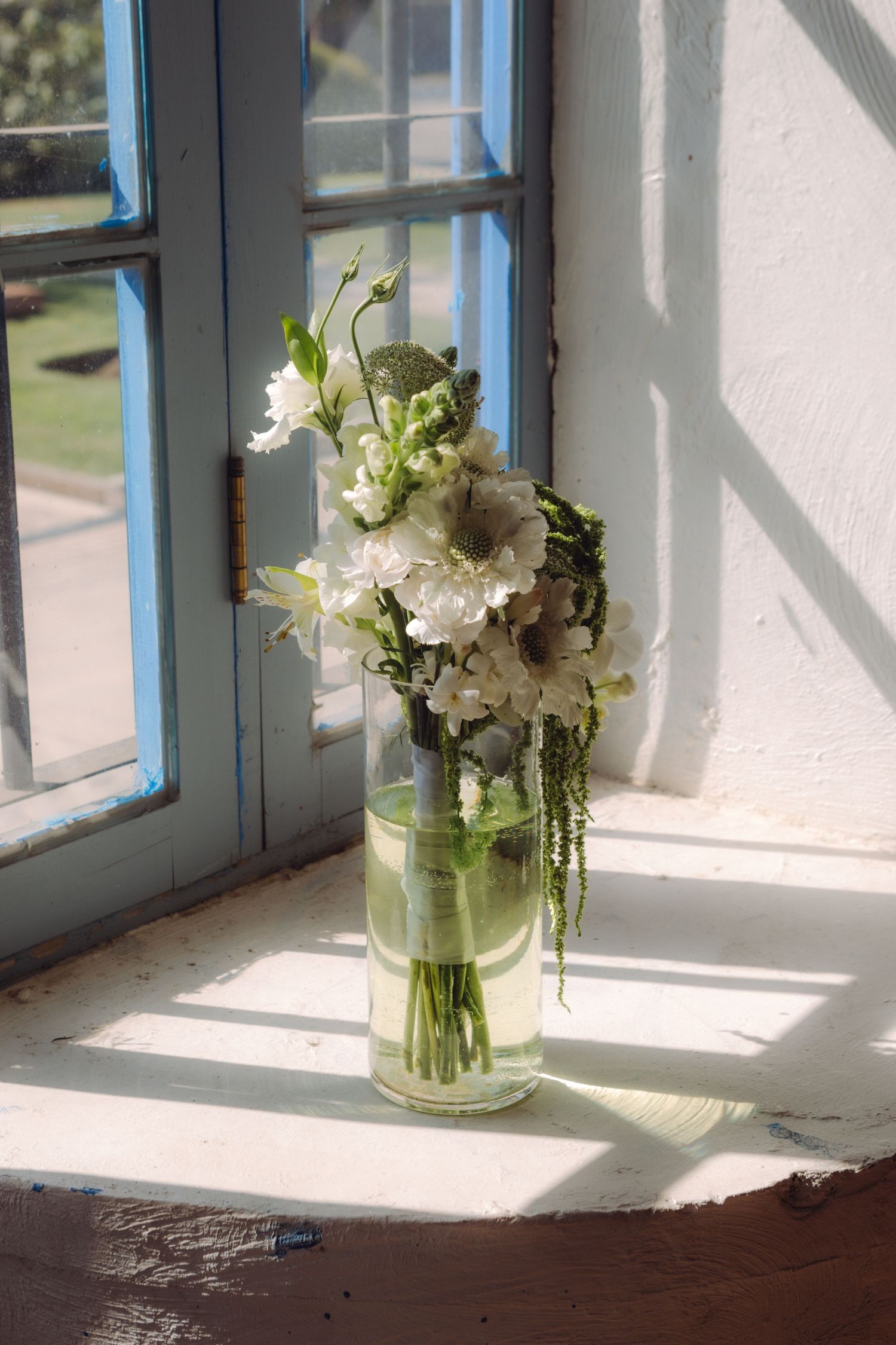 White floral bouquet in glass vase on sunlit windowsill with blue painted frame.