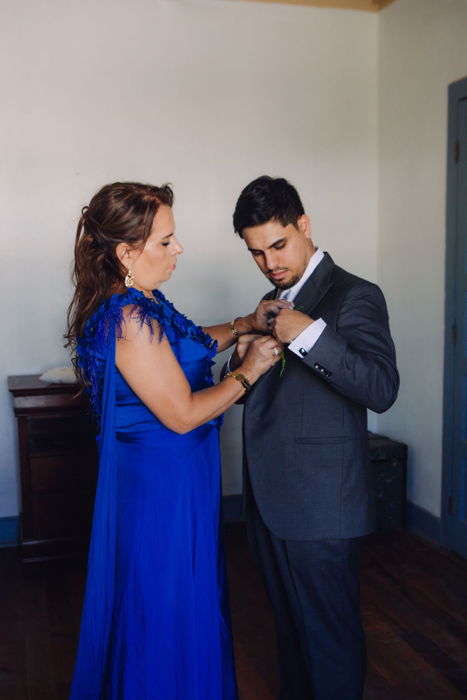 Woman in blue gown adjusting grooms suit jacket during wedding day preparations.