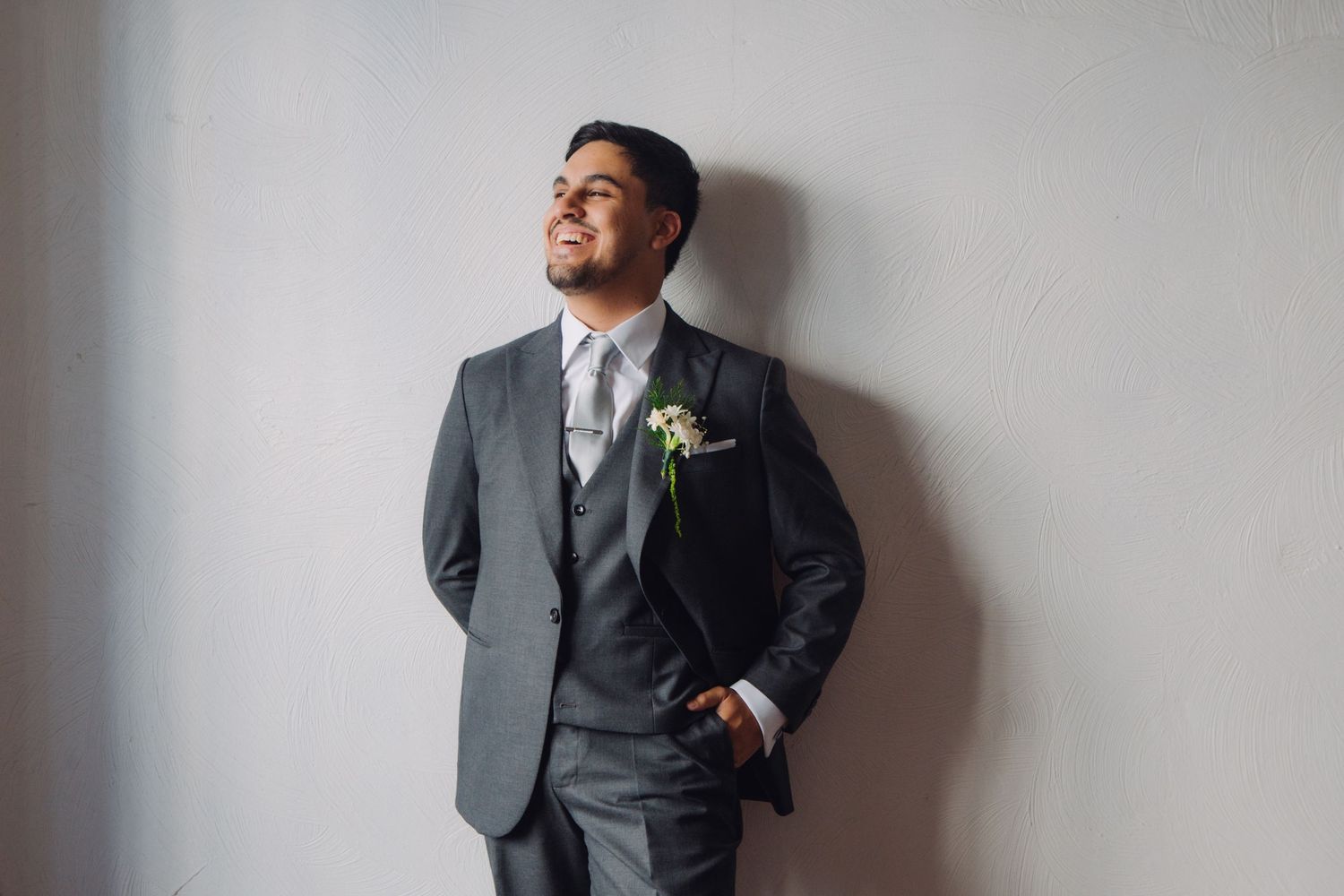 Elegant groom in gray three-piece suit with boutonniere smiling against white wall.