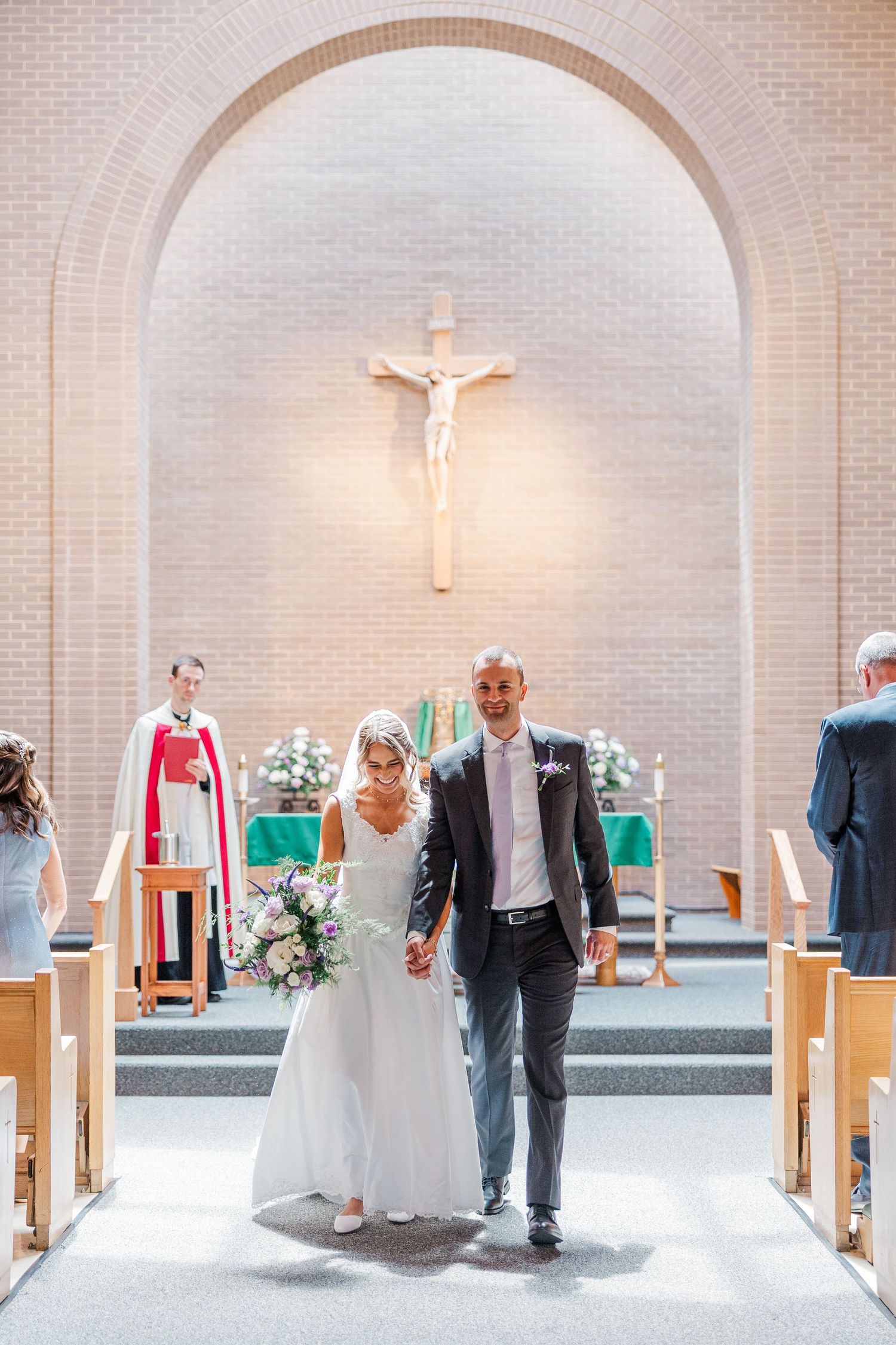 Bride and groom walk down church aisle after wedding ceremony, with priest and crucifix altar in background.