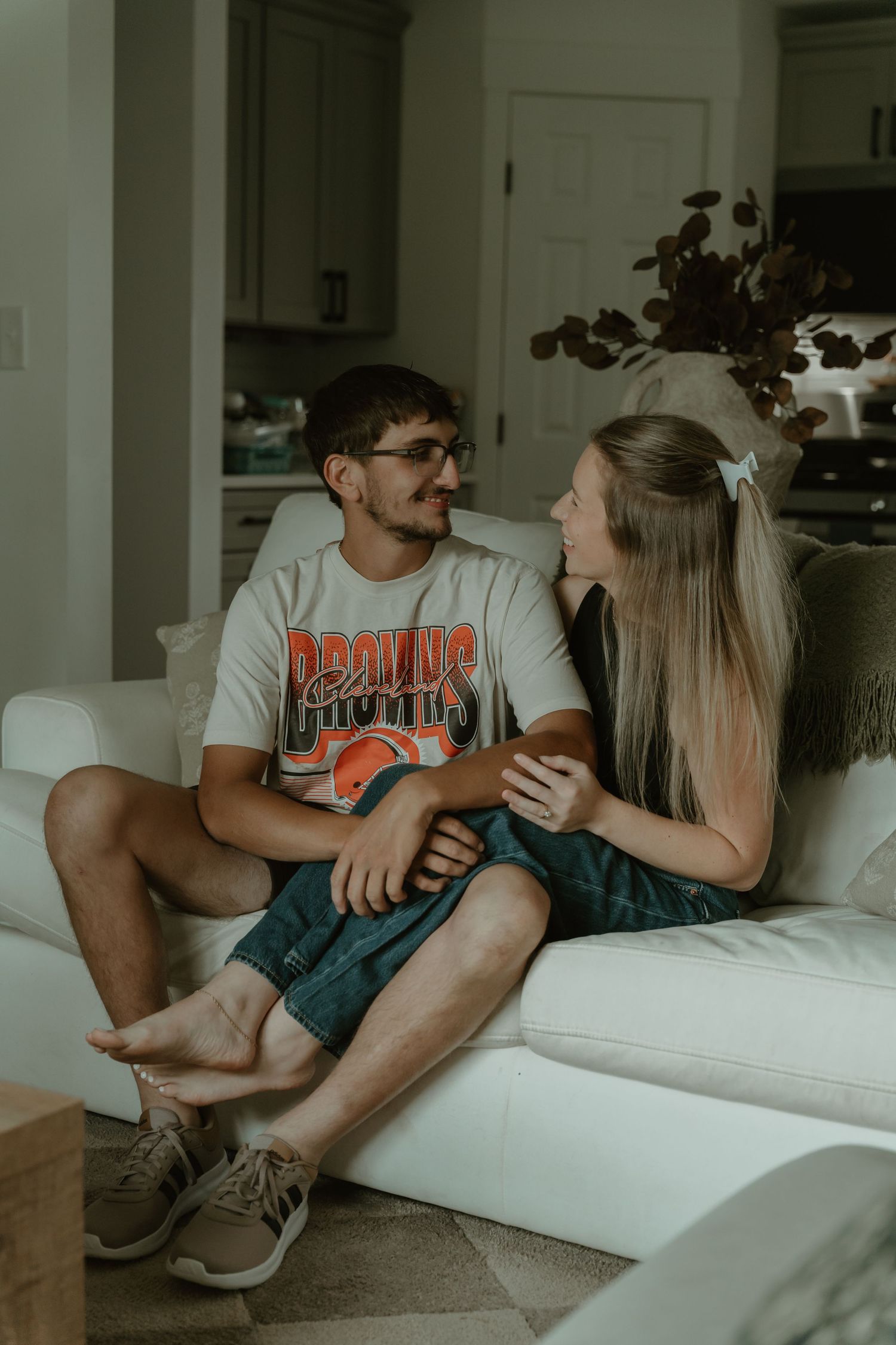 Young couple sitting together on a white couch, smiling at each other in a cozy home setting.