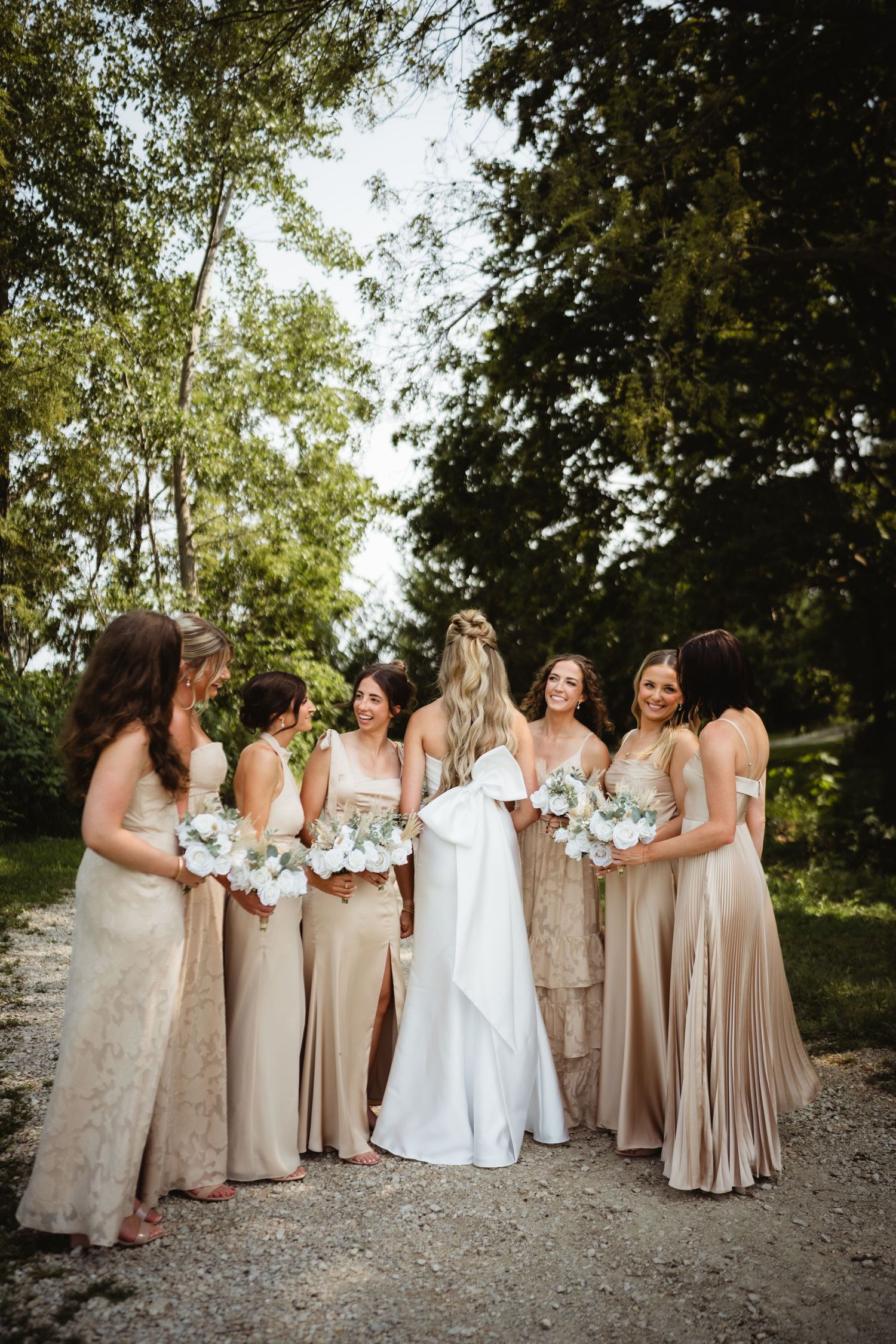 Bride in white gown with bridesmaids in champagne gold dresses holding white bouquets outdoors.