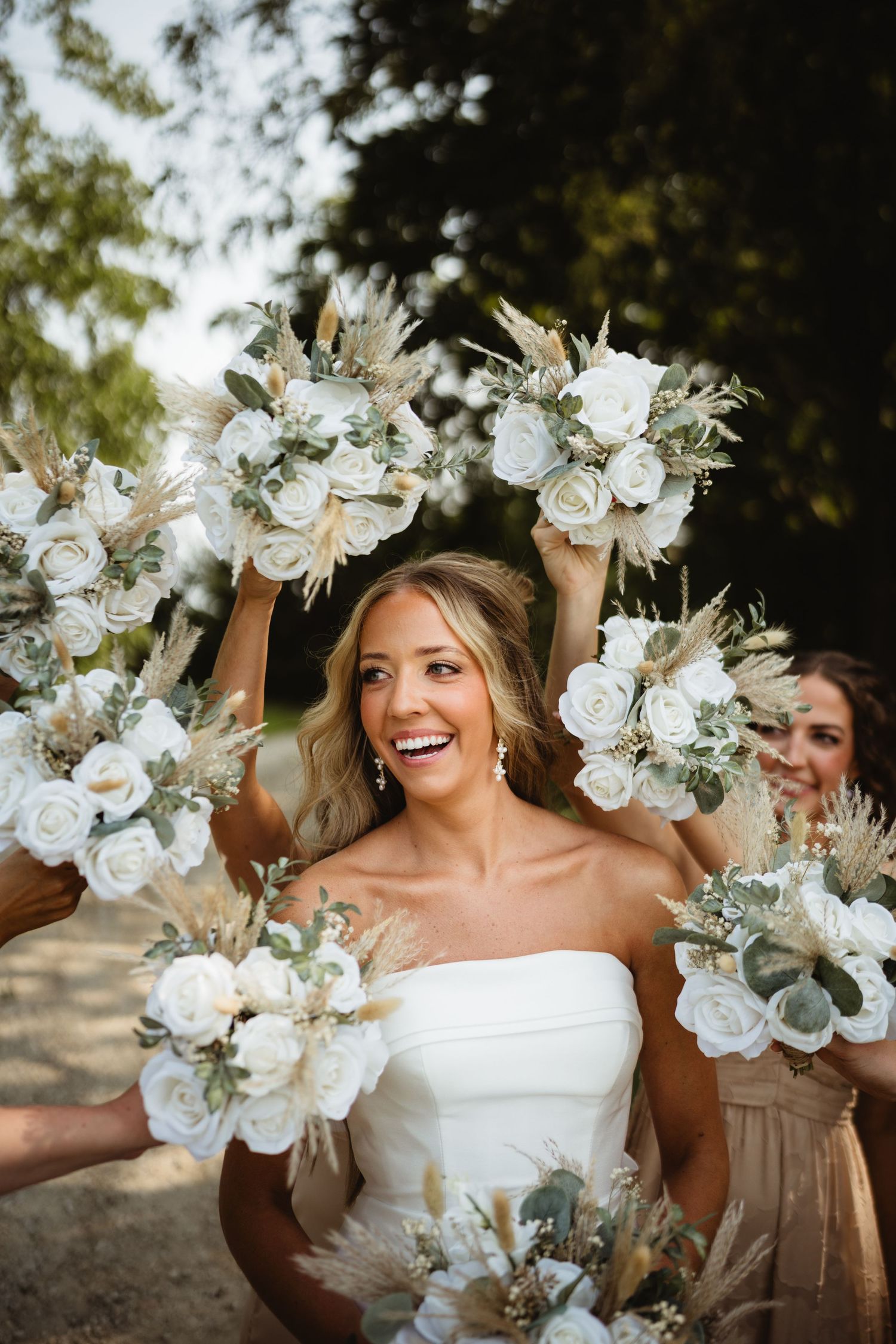 Smiling bride in white strapless gown surrounded by bridesmaids holding white rose and pampas grass bouquets outdoors.