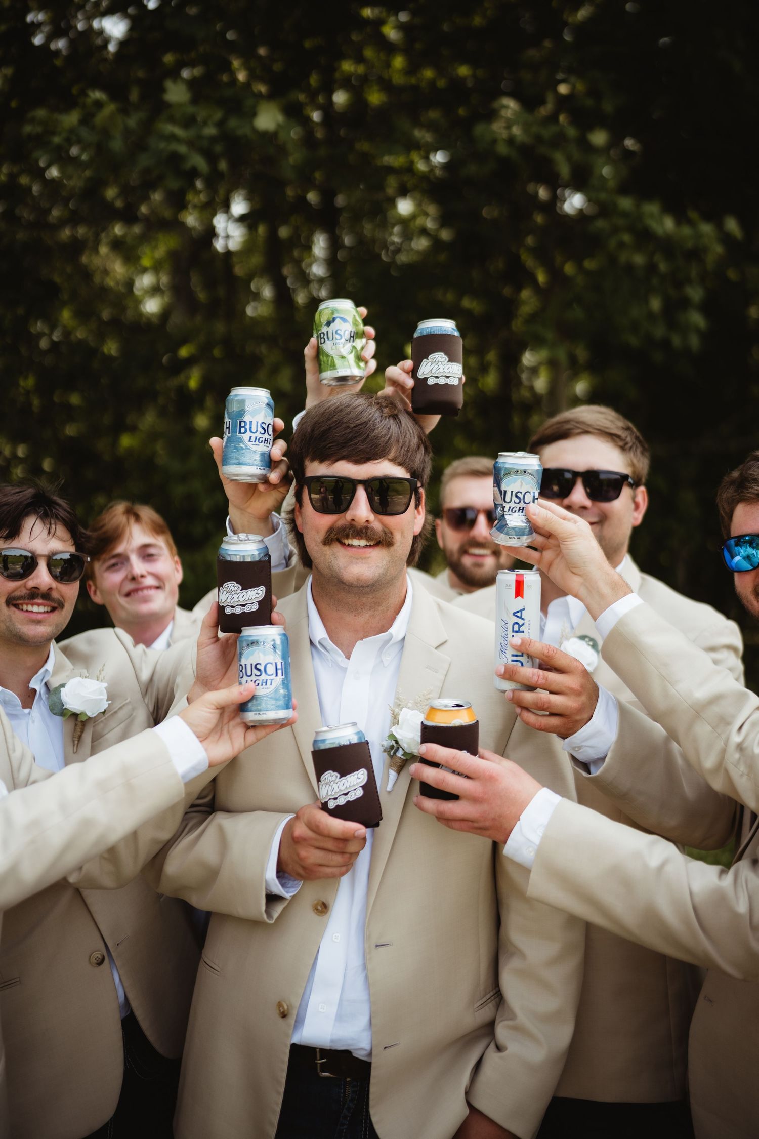 Groomsmen in tan suits and sunglasses raising beer cans in a celebratory toast outdoors at a wedding.