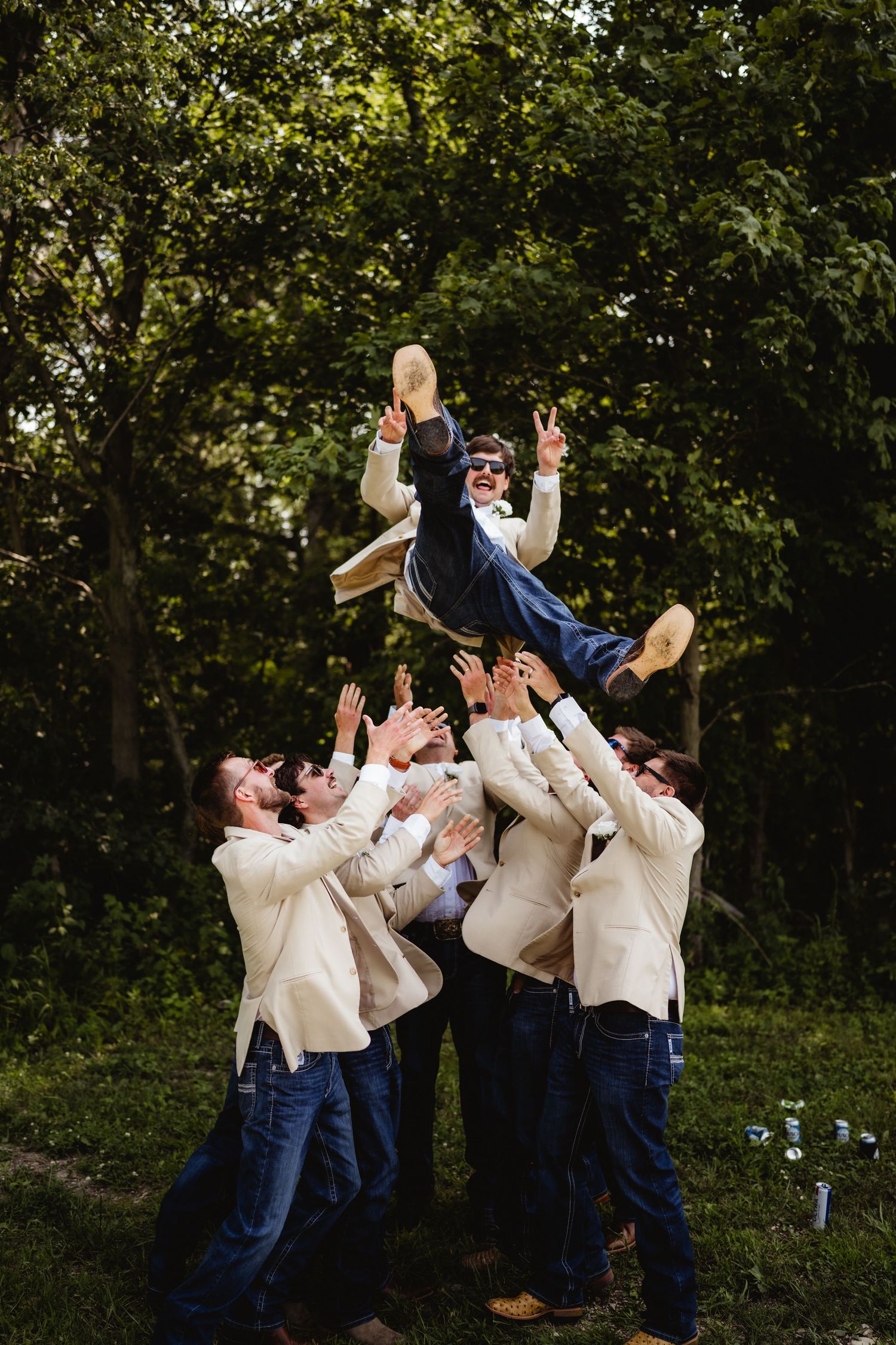 Groomsmen in cream blazers tossing groom in navy suit into the air outdoors, celebrating at a wedding.