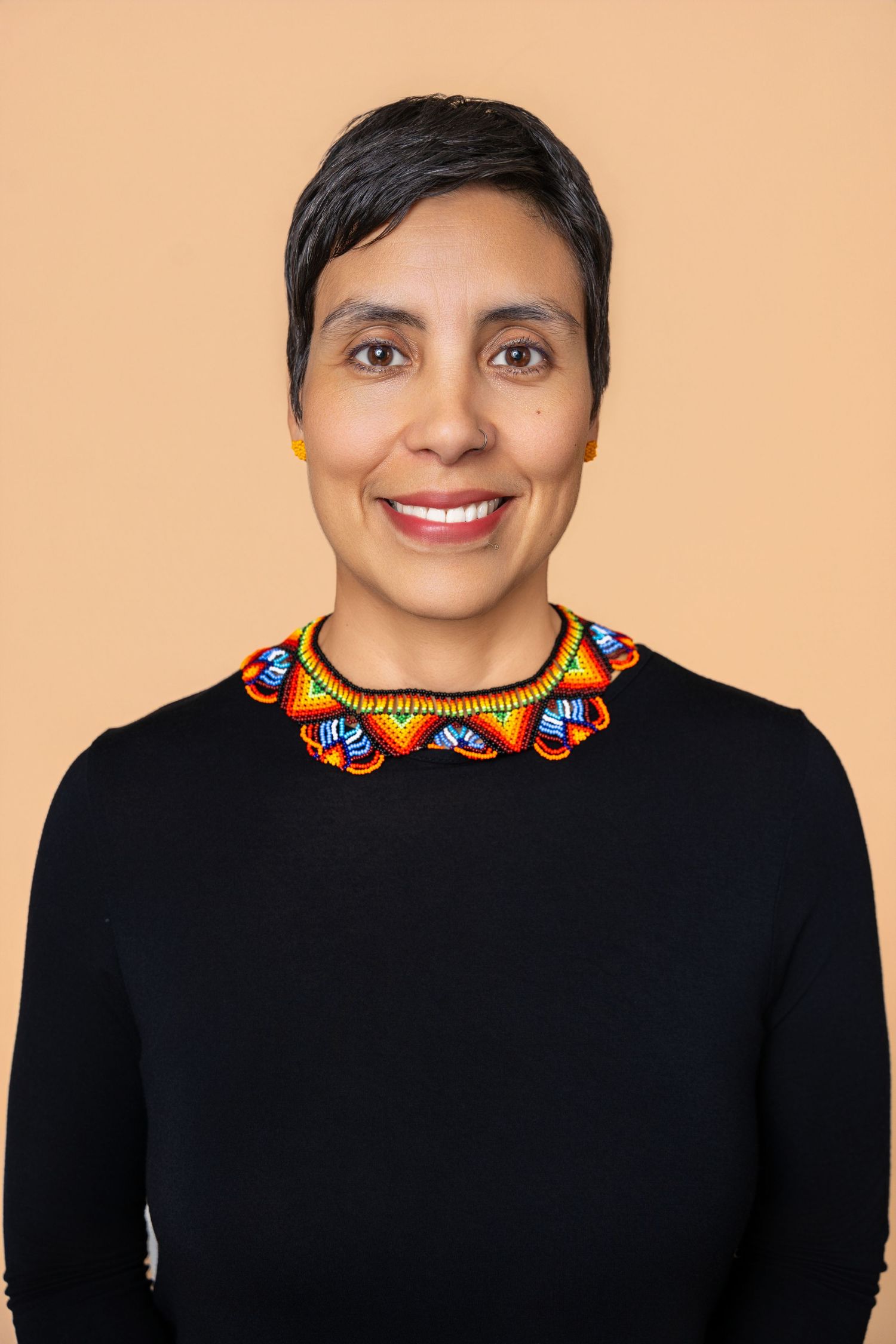 Smiling woman with short hair wearing a black top and vibrant colorful beaded necklace on beige background.