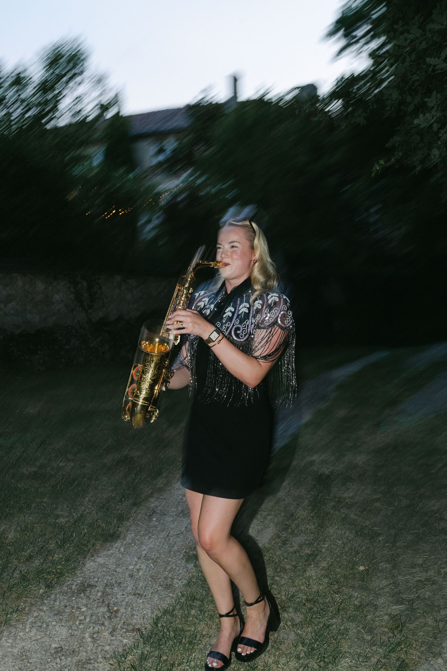 Blonde woman in black dress playing saxophone outdoors at dusk in a garden setting.