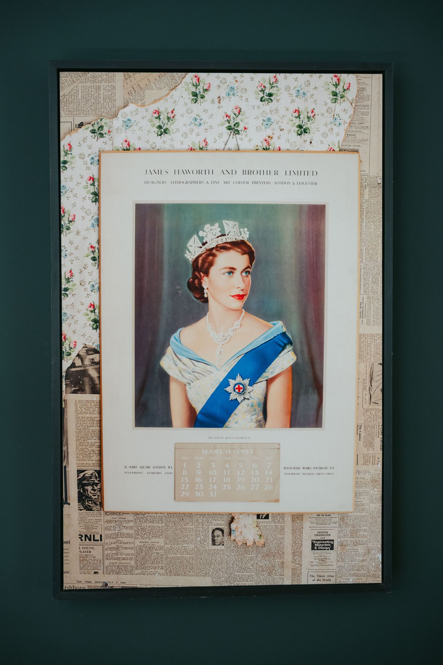 Vintage portrait of Queen Elizabeth II in tiara and blue sash, framed with newspaper, displayed on teal wall.