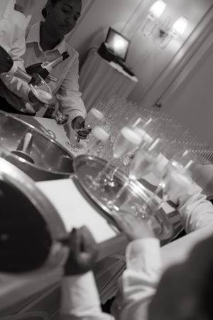 Black and white photo of catering staff pouring drinks at an elegant event bar setup with rows of glasses.