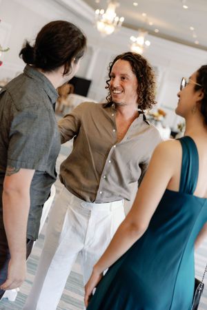 Three people socializing at an elegant indoor event, with a smiling man in a gray shirt conversing with guests.