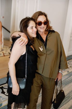 Two women posing together, one in black dress with crossbody bag, other in olive pantsuit with sunglasses and pendant necklace.