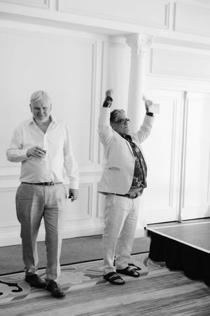 Two men celebrate at an elegant event, one raising his arm in joy while holding drinks in a classic white room.