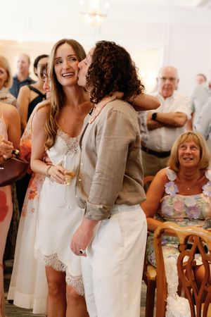 Bride in white lace dress receiving a kiss on cheek at wedding reception while holding champagne.