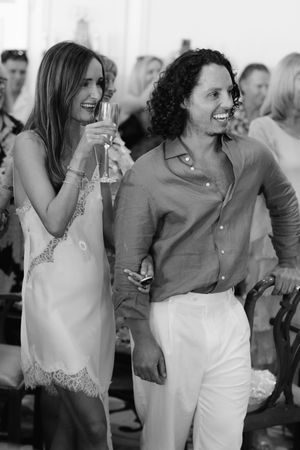 Two guests smile and laugh at a social event, woman holding champagne in a white lace dress outdoors.