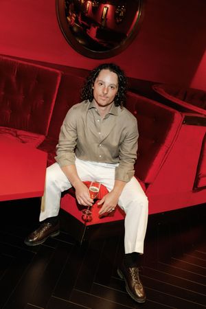 Man in beige shirt and white pants sits on red ottoman holding a drink in stylish red lounge.