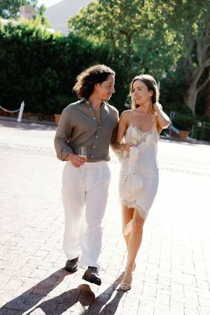 Couple walking outdoors in sunlight, man in grey shirt and white pants, woman in white slip dress, holding champagne glasses.