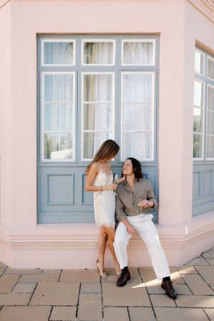 Couple in elegant attire sharing champagne outside a pastel pink building with blue French windows.