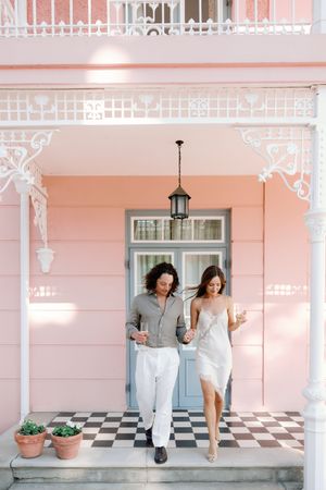 Couple walking hand-in-hand on a checkered porch of a pink Victorian building with white ornate trim.
