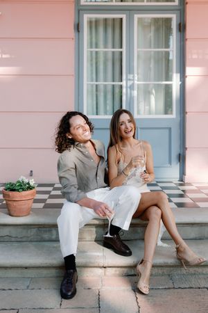Happy couple sitting on checkered steps of a pink building, holding champagne glasses and smiling.