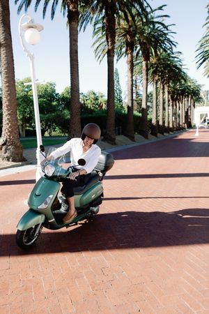 A smiling woman wearing a helmet rides a green scooter down a palm tree-lined brick boulevard on a sunny day.