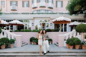 Two women posing together in front of a elegant pink hotel with white umbrellas and lush potted plants.