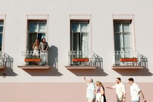 People walking past a building with balconies while two people watch from above.