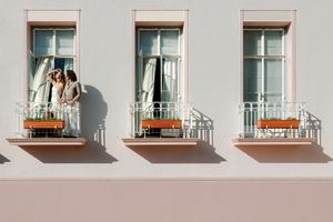 Two people stand on a decorative balcony of a pastel pink building with three French windows and ornate railings.