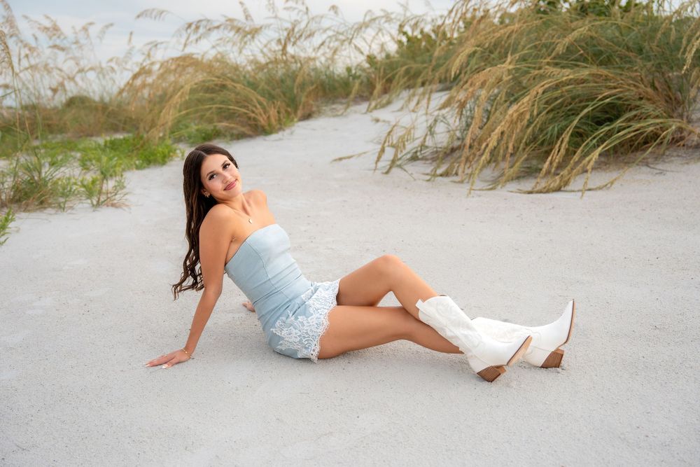 A young woman in a blue strapless dress and white cowboy boots sits on sandy beach dunes.