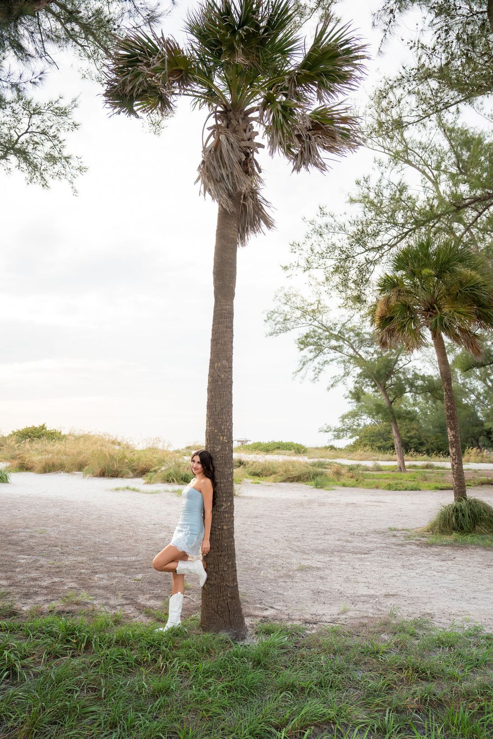 Brunette woman in blue dress and white boots posing against a tall palm tree at a sandy coastal park.