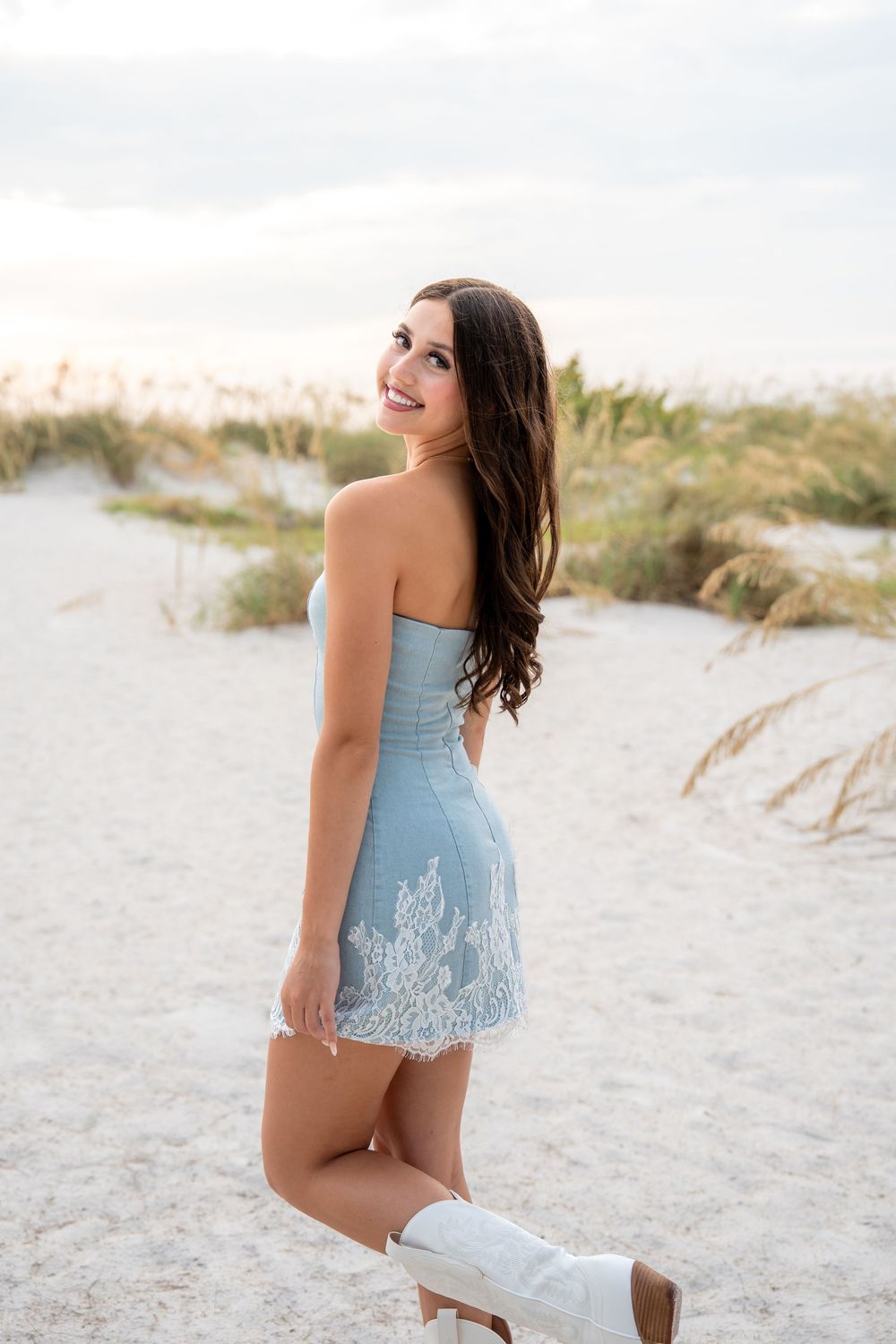 Young woman in strapless light blue lace mini dress and white boots posing on a white sand beach.